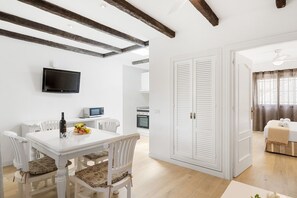 Zona pranzo con tavolo e sedie bianche, televisione a parete e cucina sullo sfondo.