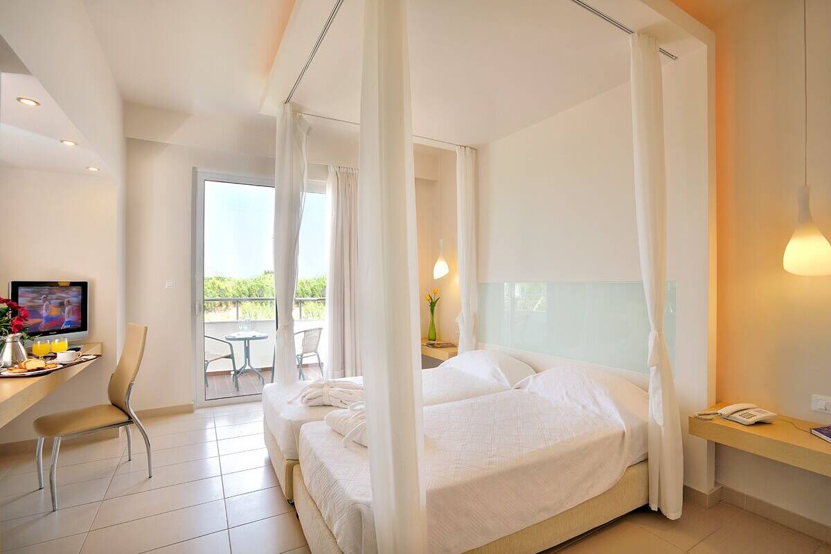 A hotel room with a bed, a desk with a chair, a TV, and a balcony with a table and chairs.