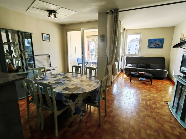 A living room with a sofa, a dining table with chairs, a television, and a window with curtains. - Spacious T3 in Port Center, Residence with Pool, Cap d'Agde (Cap d'Agde)
