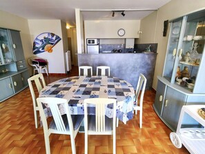 A dining area with a round table covered by a patterned tablecloth, surrounded by white chairs. A kitchen with a microwave and stove is visible in the background. - Spacious T3 in Port Center, Residence with Pool, Cap d'Agde (Cap d'Agde)