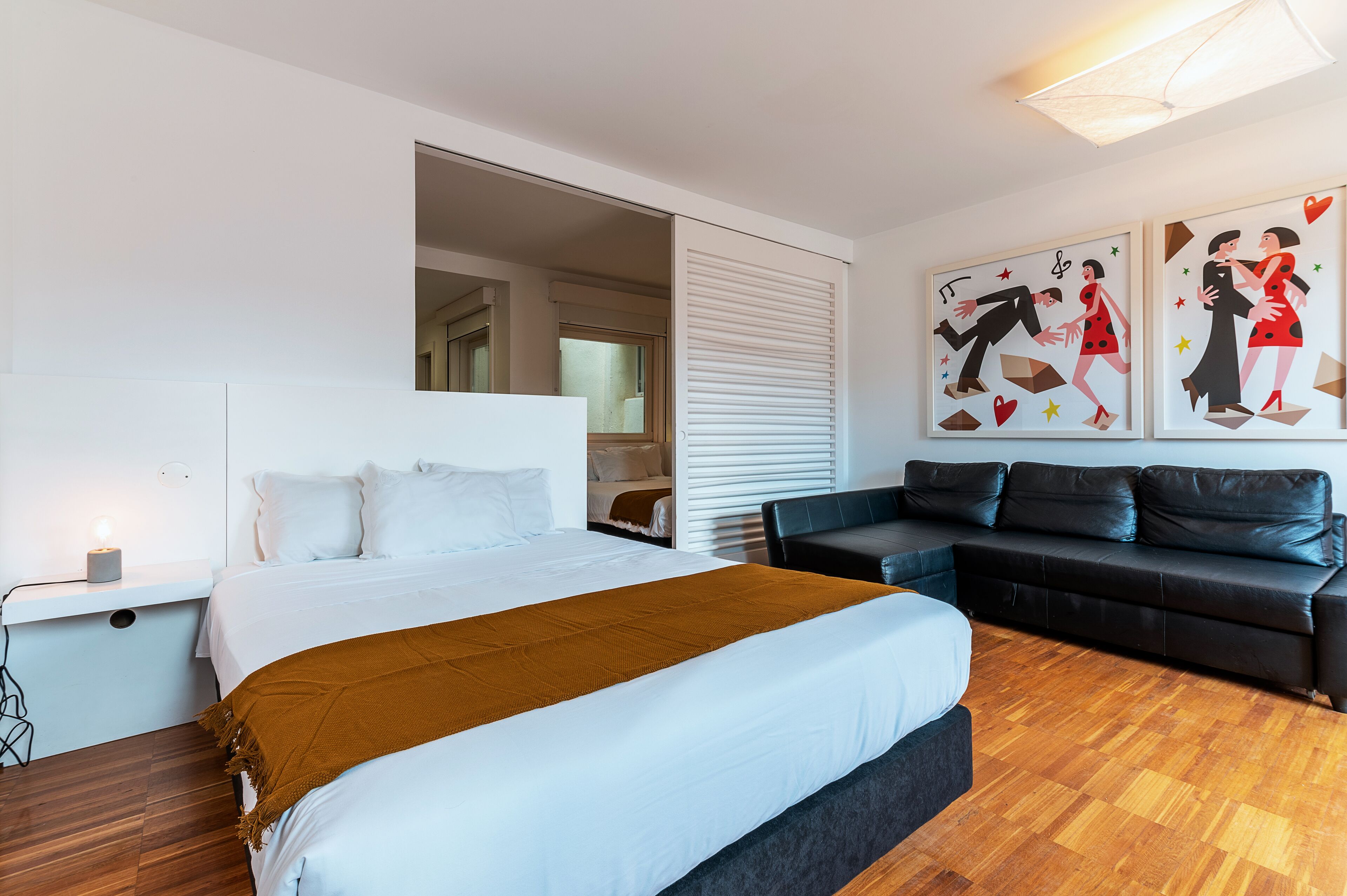 A modern hotel room with a large bed, a black leather sofa, and two wall-mounted artworks depicting dancers.