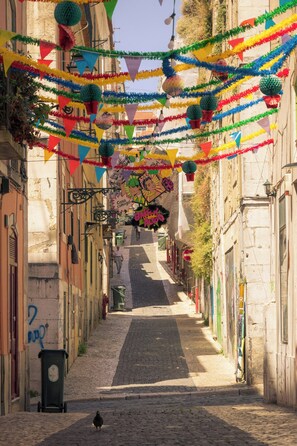 A narrow cobblestone street with buildings on either side, decorated with colorful flags and hanging decorations. - Portas do Bairro Alto (Lisbon)