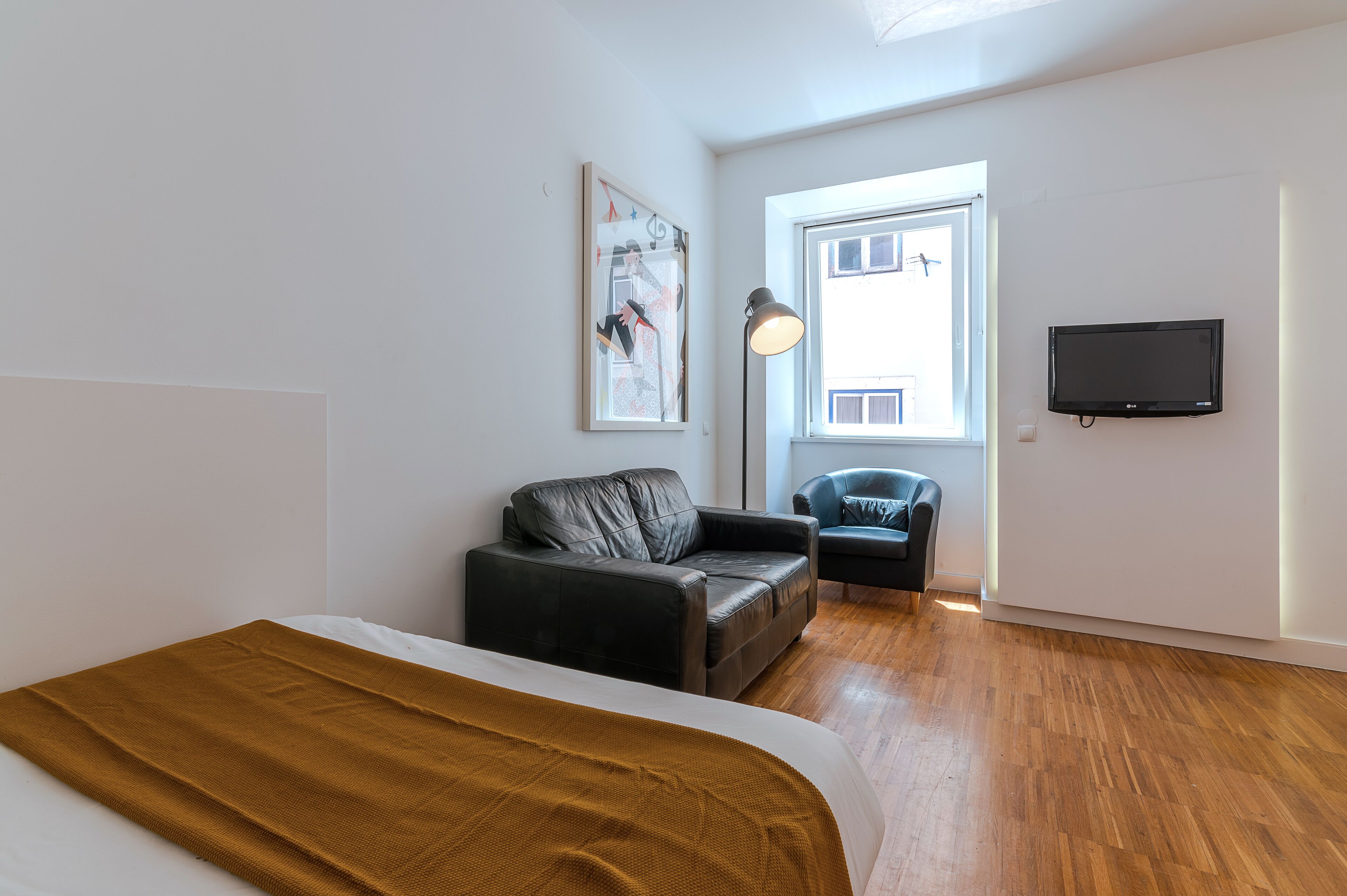 Et moderne hotelværelse med seng, sort lædersofa, en mindre lænestol, et fladskærms-tv monteret på væggen og et stort vindue, der giver naturligt lys.