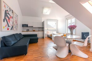 A modern living room with a black sofa, white chairs, a dining table with a vase of flowers, and a kitchen area with white cabinets and a microwave. - Portas do Bairro Alto (Lisbon)