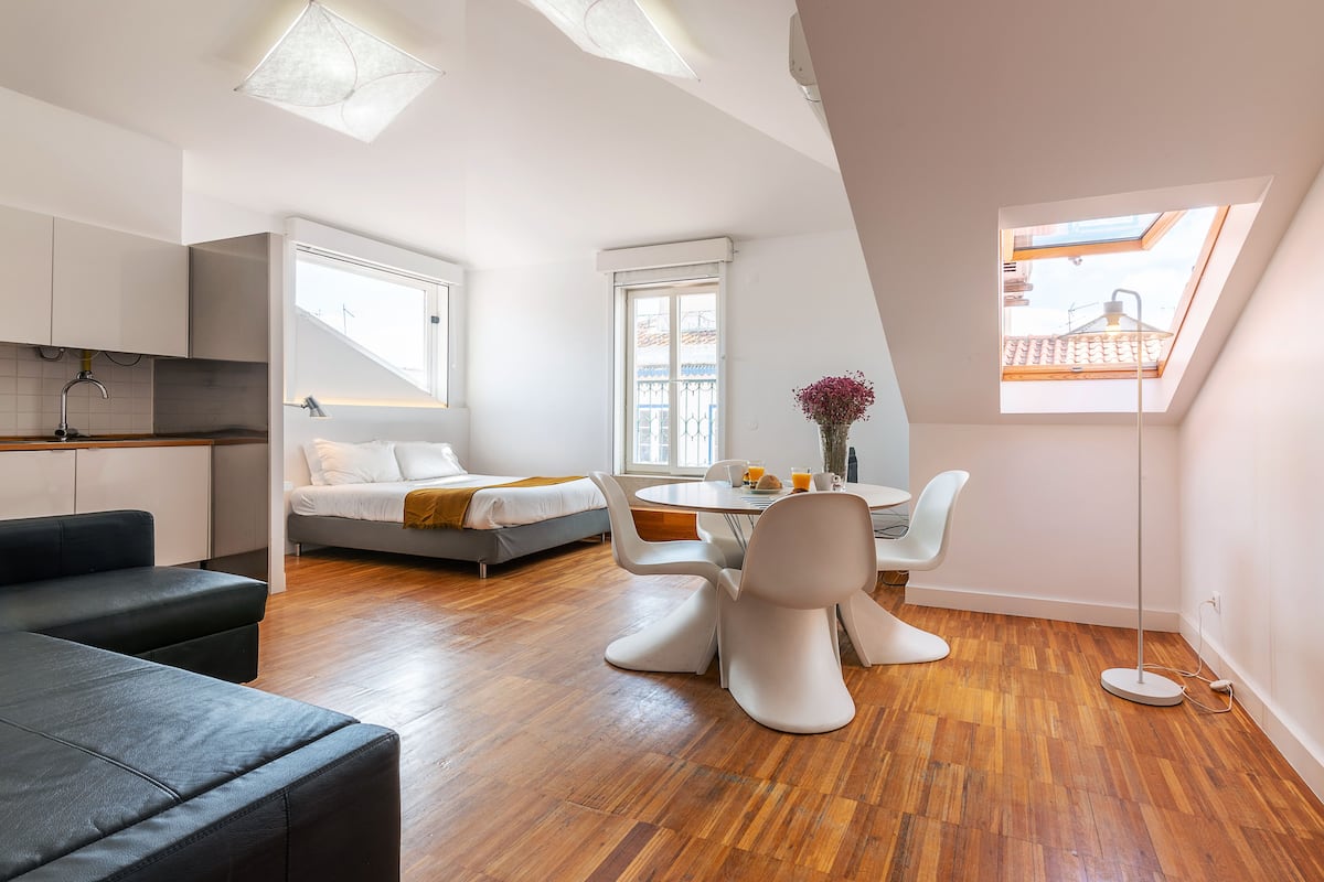 A modern loft-style room with a dining area, a sofa, a bed, and a skylight.