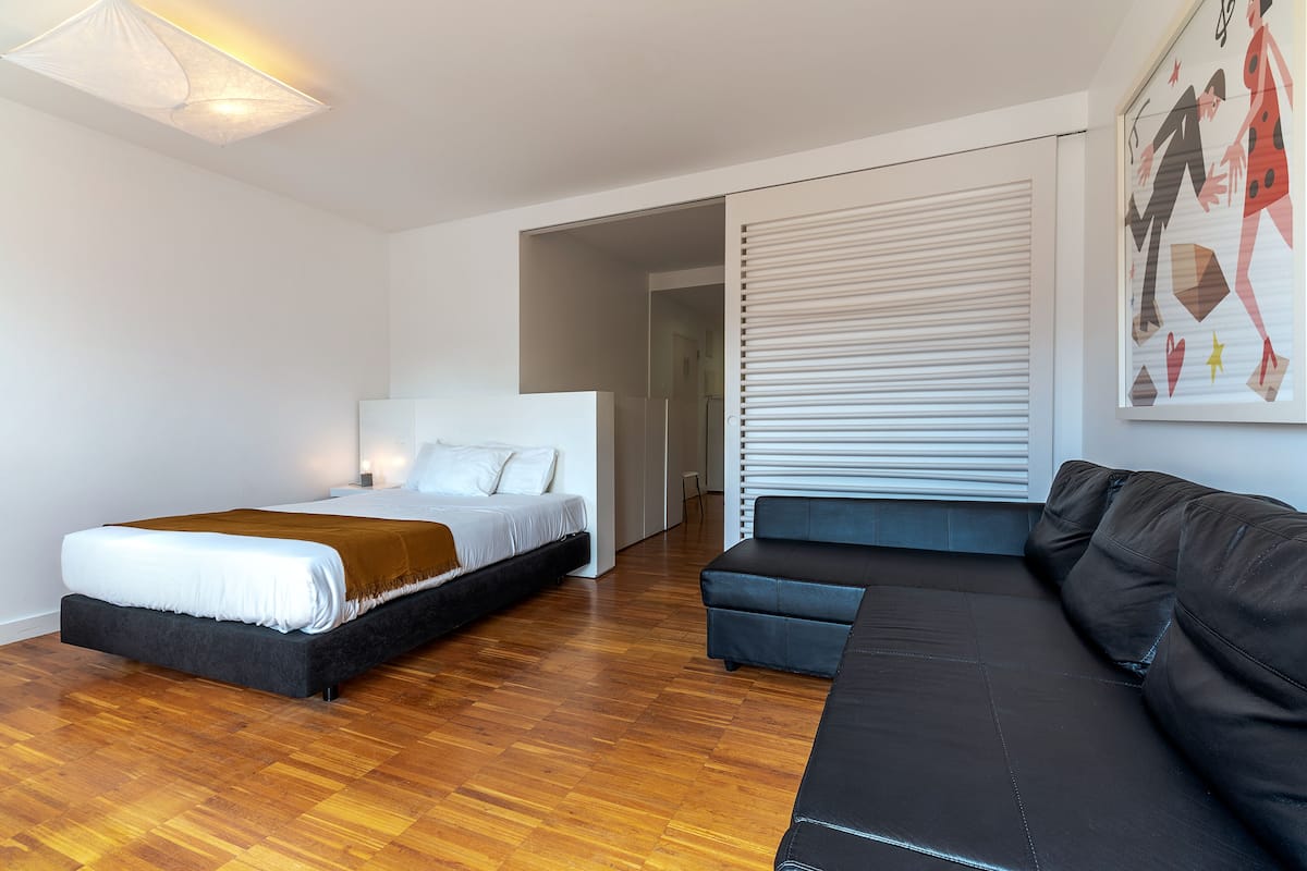 A modern bedroom with a bed, a sofa, and a large window with blinds.