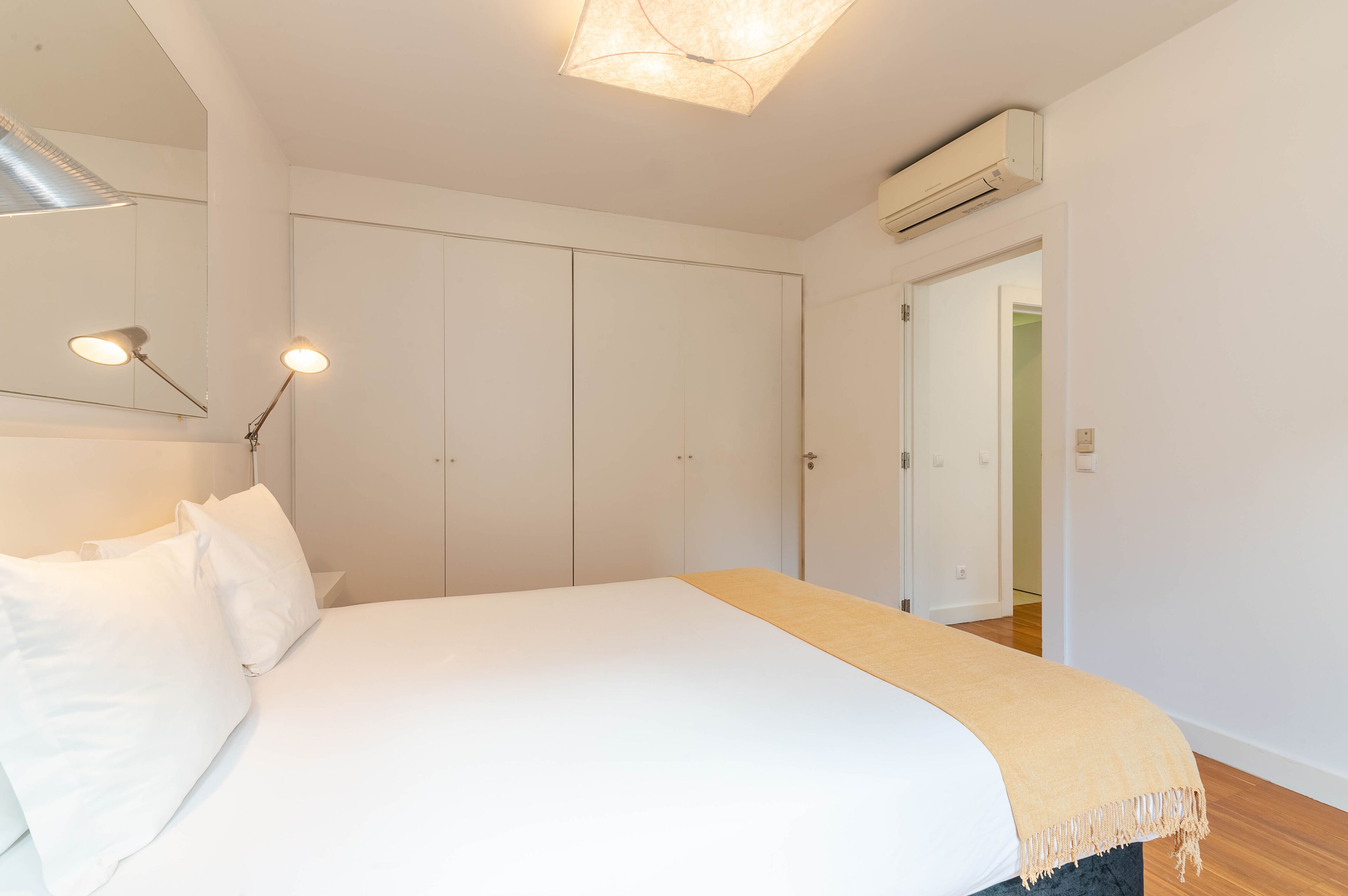 Quarto com cama, paredes brancas, porta e aparelho de ar condicionado.