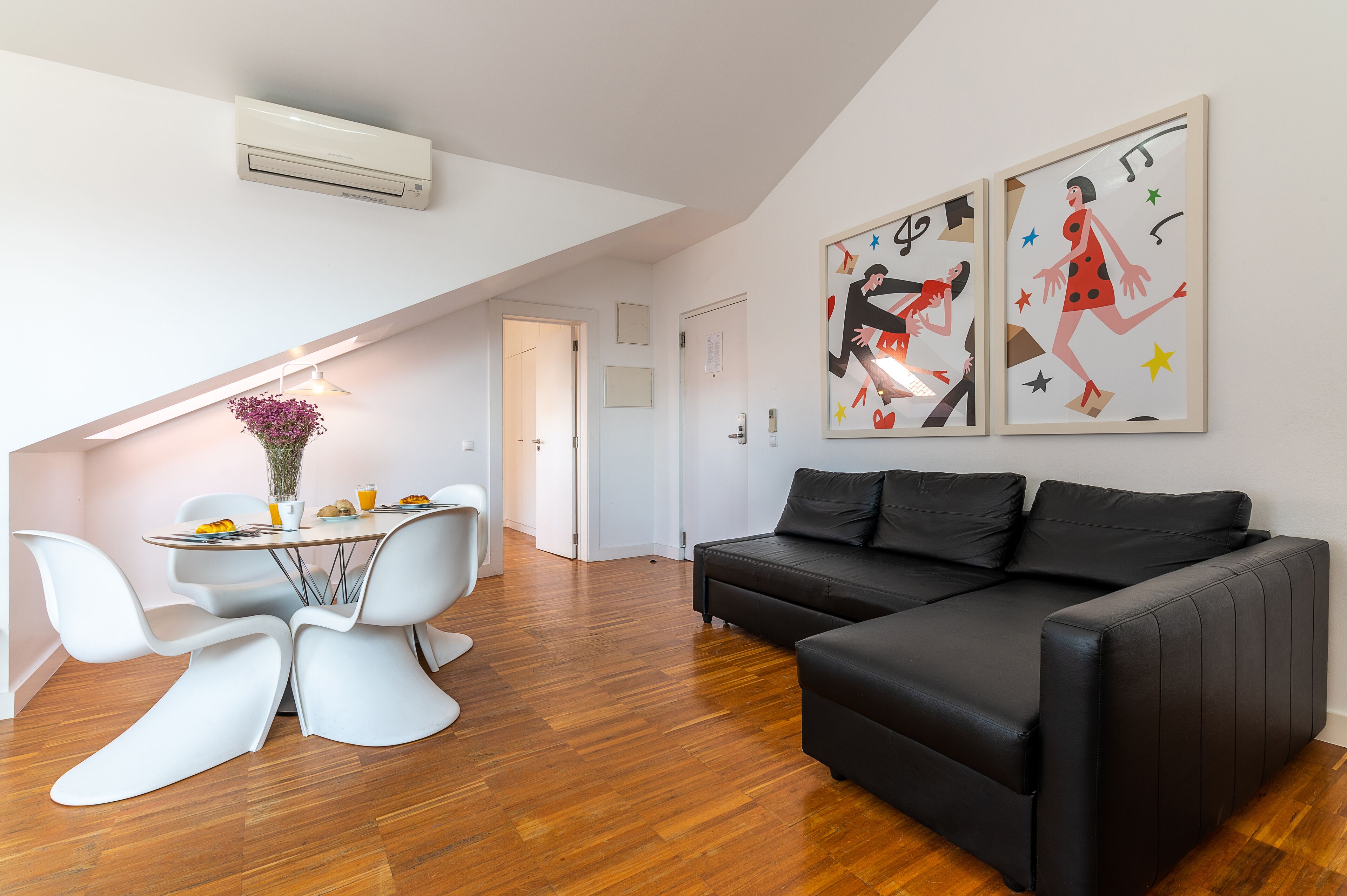 A modern living space with a dining area, a black leather sofa, and two framed artworks on the wall.