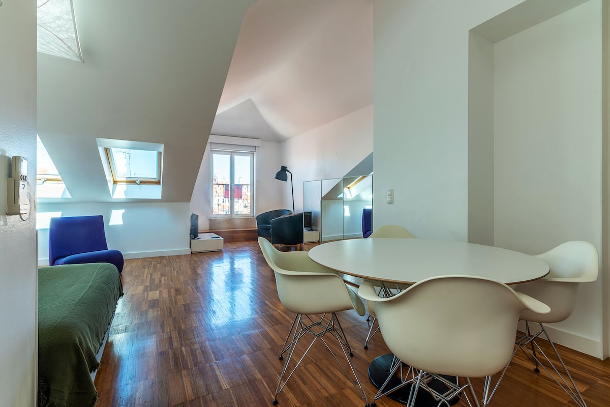 A modern room with a wooden floor, a round table, and several chairs. There is a bed with a green blanket, a blue chair, and a window with a view of buildings.