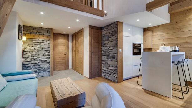 Un vaste espace de vie agrémenté d’un mur en pierre, d’un plafond en bois et d’une cuisine équipée d’un réfrigérateur et de tabourets de bar.