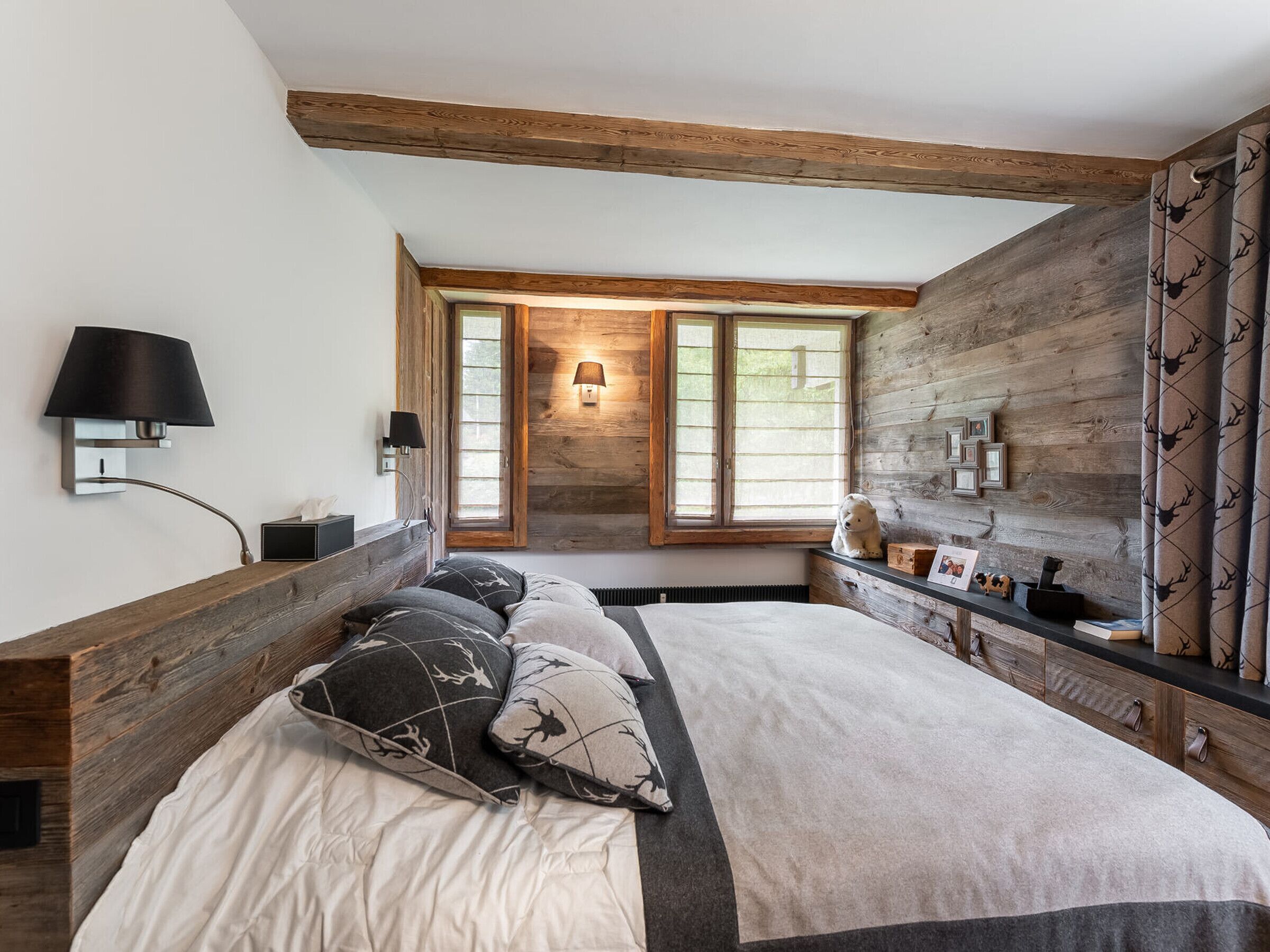 A bedroom with a large bed, wooden beams, a window with a view, and a bedside table with a lamp.