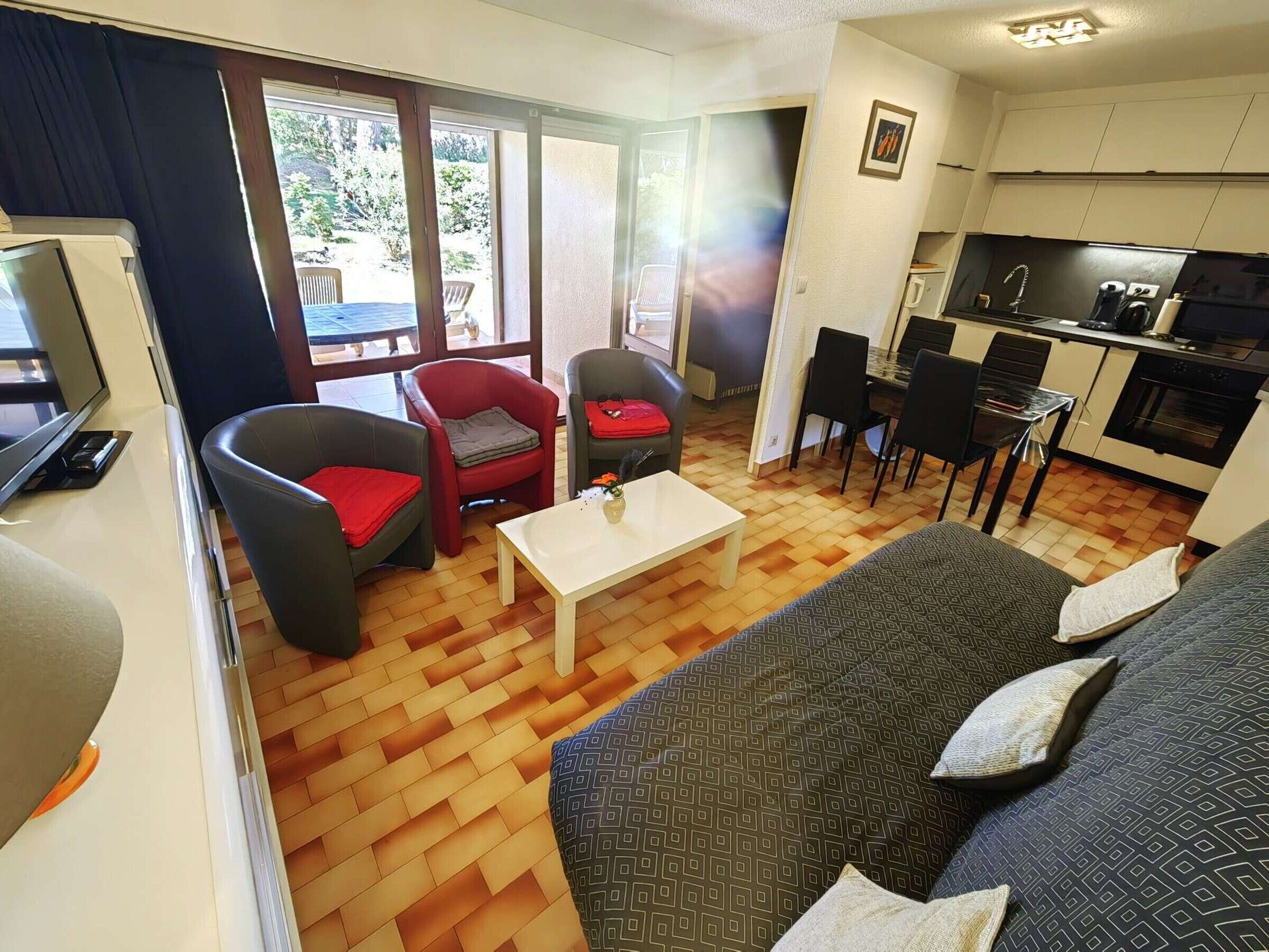 A living room with a sofa, chairs, a coffee table, and a TV. There is a kitchen area with a dining table and chairs.