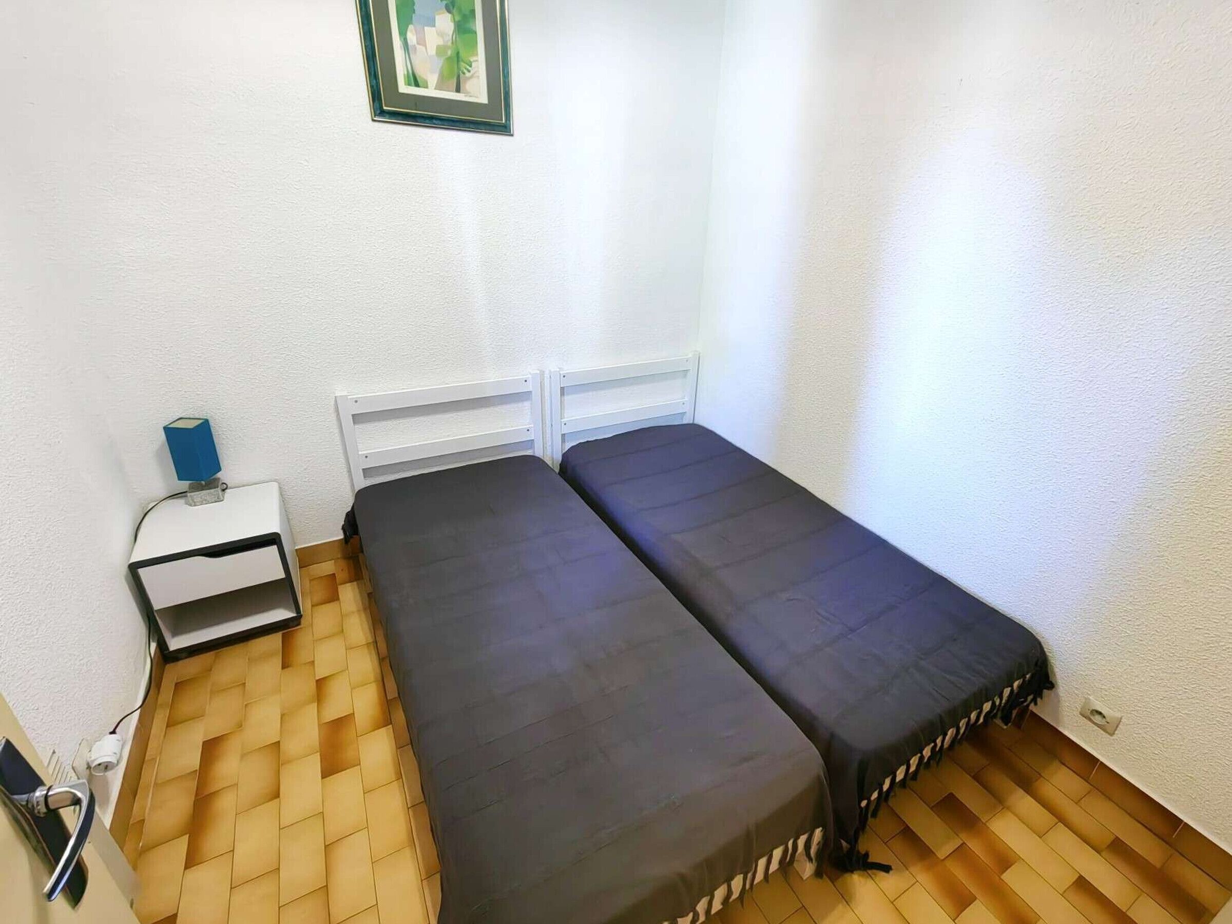 Ett litet rum med två enkelsängar, ett nattduksbord med en lampa, ett eluttag och en inramad tavla på väggen.