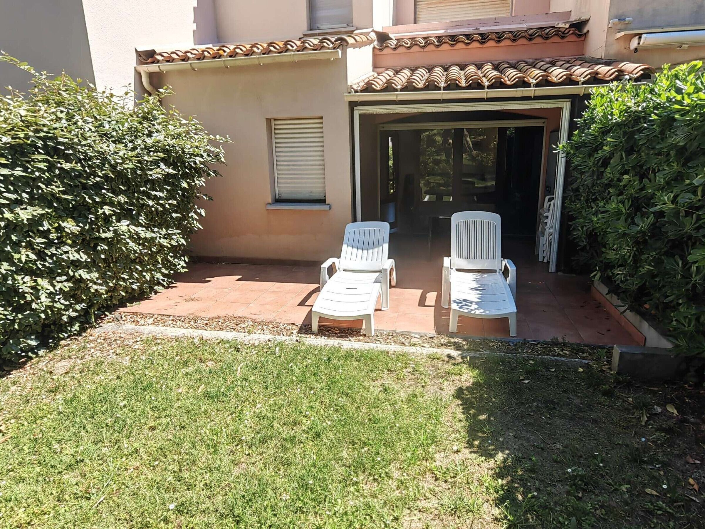 An outdoor seating area with two white lounge chairs and a table, surrounded by a well-maintained lawn and a large bush.