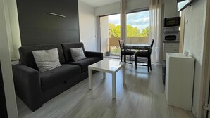 Een moderne woonkamer met een zwarte bank, een witte salontafel en een eethoek met een tafel en stoelen. Er is een keuken met een koelkast en een magnetron.