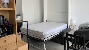Een kleine kamer met een bed, een bureau, een stoel, een kledingkast en een plank met een lamp en een koffiezetapparaat.