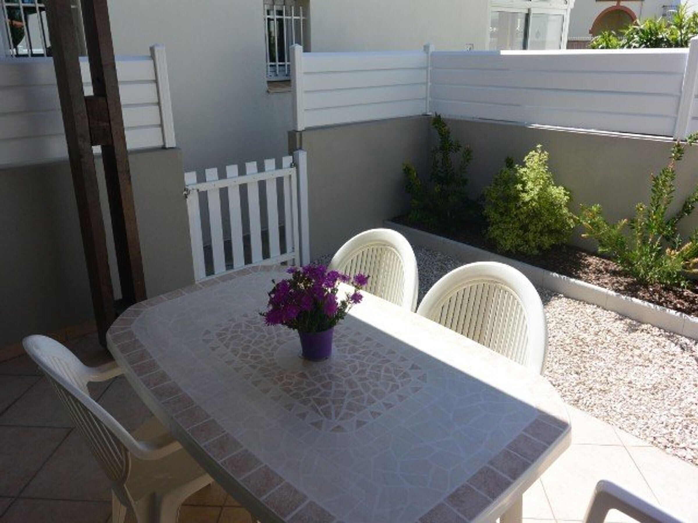 A patio with a table and chairs, a small garden, and a white fence.