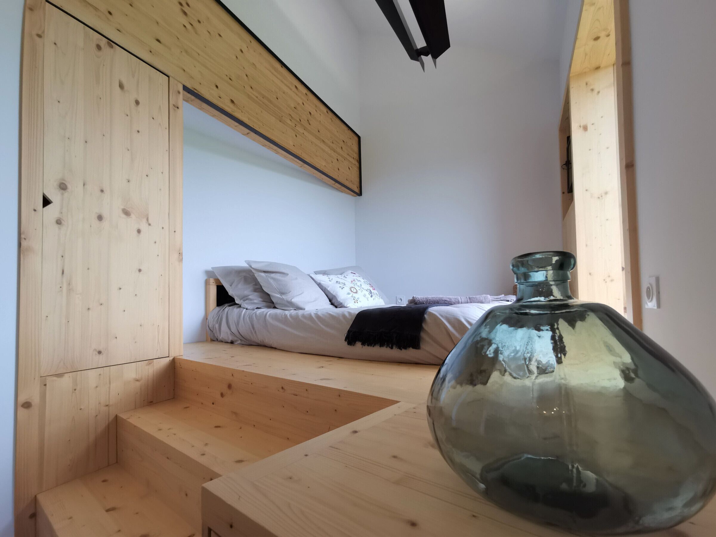 Une pièce comprenant un lit, un grand vase en verre et un escalier en bois menant à une mezzanine.