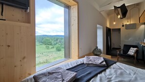 A bedroom with a large window, a bed with folded blankets, a chair, and a hanging light fixture. - Nature Retreat: Cozy Cottage with Panoramic Views, Perfect for Family & Friends Near Lyon & Clermont-Ferrand (Saint-Victor-sur-Arlanc)