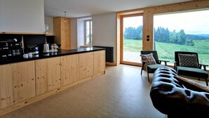 A modern kitchen with wooden cabinets and a black countertop, a cozy seating area with a leather sofa and chairs, and a large sliding glass door leading to a scenic outdoor view. - Nature Retreat: Cozy Cottage with Panoramic Views, Perfect for Family & Friends Near Lyon & Clermont-Ferrand (Saint-Victor-sur-Arlanc)