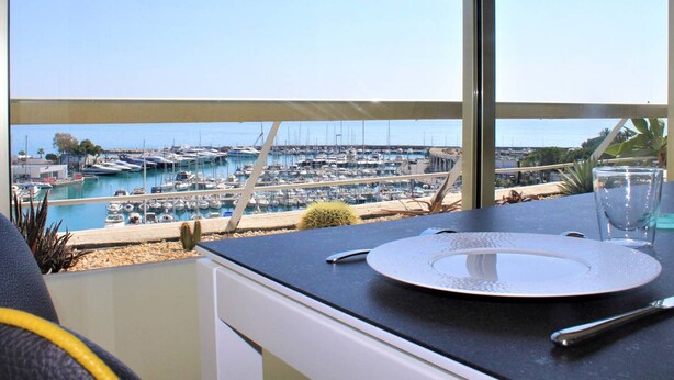 Een eettafel met een bord, een glas en een lepel, met uitzicht op een jachthaven met boten.