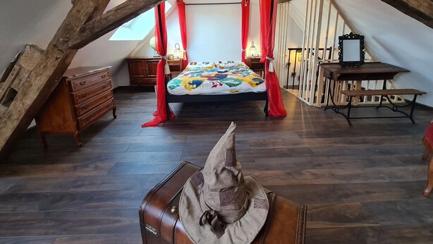 Une chambre avec un plafond en pente, un lit recouvert d’une courtepointe aux couleurs vives, des rideaux rouges, un bureau en bois et une valise.
