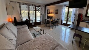 A modern living room with a sofa, dining table, and chairs. There is a kitchen area with a TV and a window with curtains. - Spacious Home with Pool, Tennis, and Garden - Sleeps 10 (Vendres)