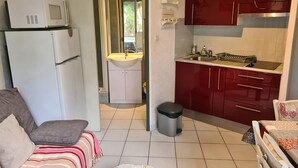Un espace de vie compact comprenant un coin cuisine, un canapé, une petite poubelle et une salle de bain visible à travers une porte ouverte.