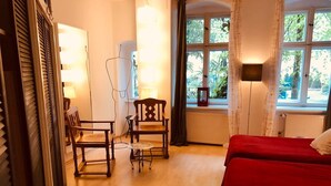 Une pièce avec un plancher en bois, deux chaises en bois, une petite table, un lit rouge, une fenêtre donnant sur un paysage verdoyant et un radiateur.