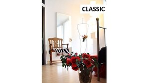 Une pièce avec un plancher en bois, une chaise, un vase rempli de fleurs rouges et un miroir accroché au mur.