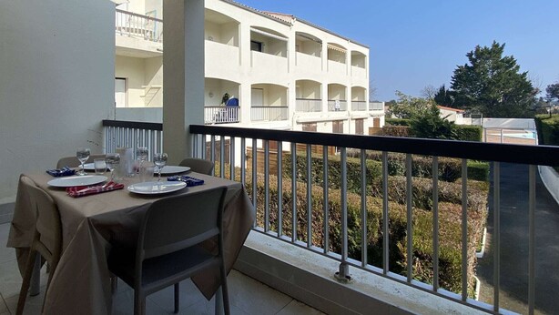 A balcony with a table set for dining, overlooking a building with multiple balconies and a clear sky.