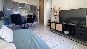 A modern living room with a large flat-screen TV, a sofa, a dining area with chairs, and a kitchen in the background.