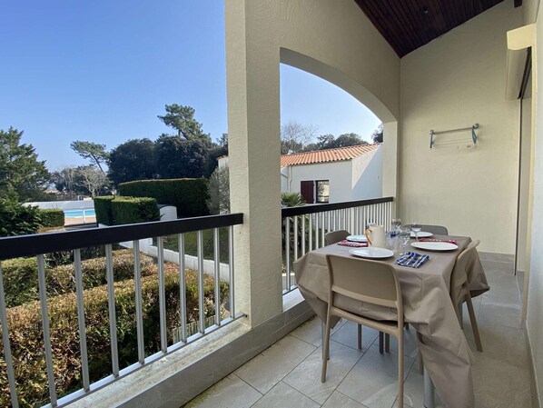 Un balcon avec une table dressée pour deux personnes, une rambarde en métal et une vue sur une piscine et des arbres.