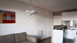 A modern living room with a grey sofa, a wooden dining table, and a kitchen area with white cabinets and a stainless steel stove.
