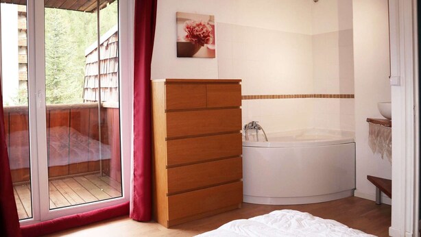 A bedroom with a wooden dresser, a white bed, a bathtub, and a sliding glass door leading to a balcony.