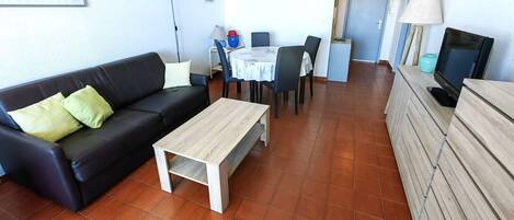 A living room with a black sofa, a wooden coffee table, a dining table with chairs, and a TV on a wooden cabinet.