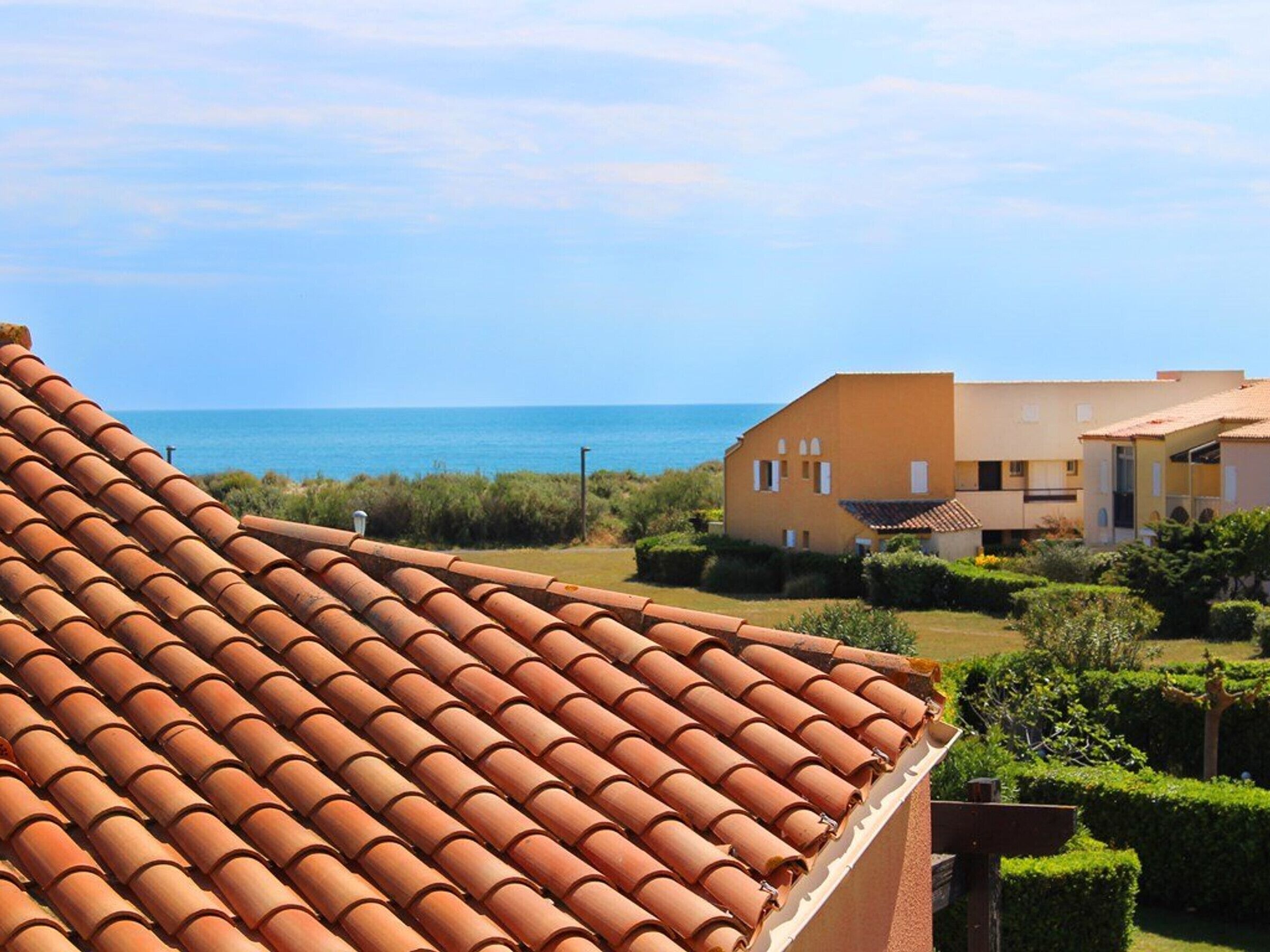 A coastal building with terracotta roof tiles, a clear view of the sea, and well-maintained greenery.