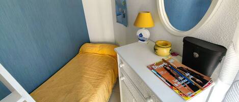 A small, neatly arranged bedroom with a single bed, a bedside table with a lamp, a mirror, and a dresser with a magazine on top.