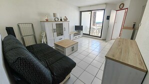Ein Wohnzimmer mit einer Couch, einem Fernseher, einem Schrank und einer Glastür zum Balkon.