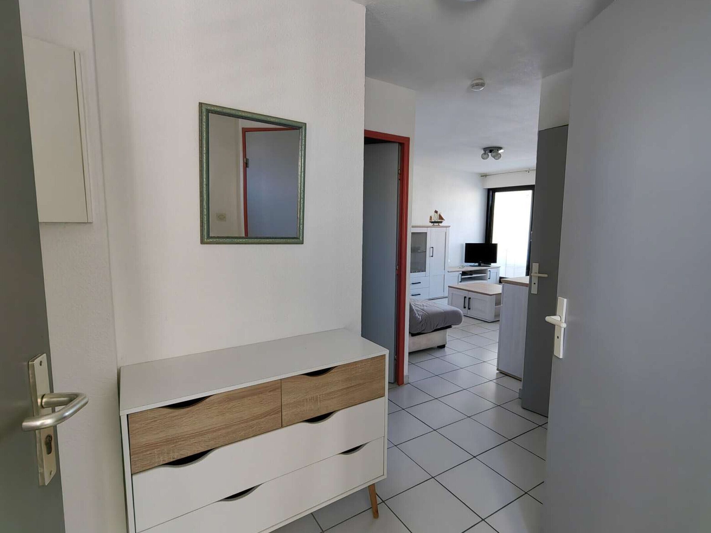 Une pièce moderne et bien éclairée, avec une commode blanche, un miroir et un téléviseur à l’arrière-plan.