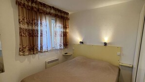 Une chambre à coucher avec un lit, une fenêtre avec des rideaux, une table de chevet et un luminaire fixé au mur.