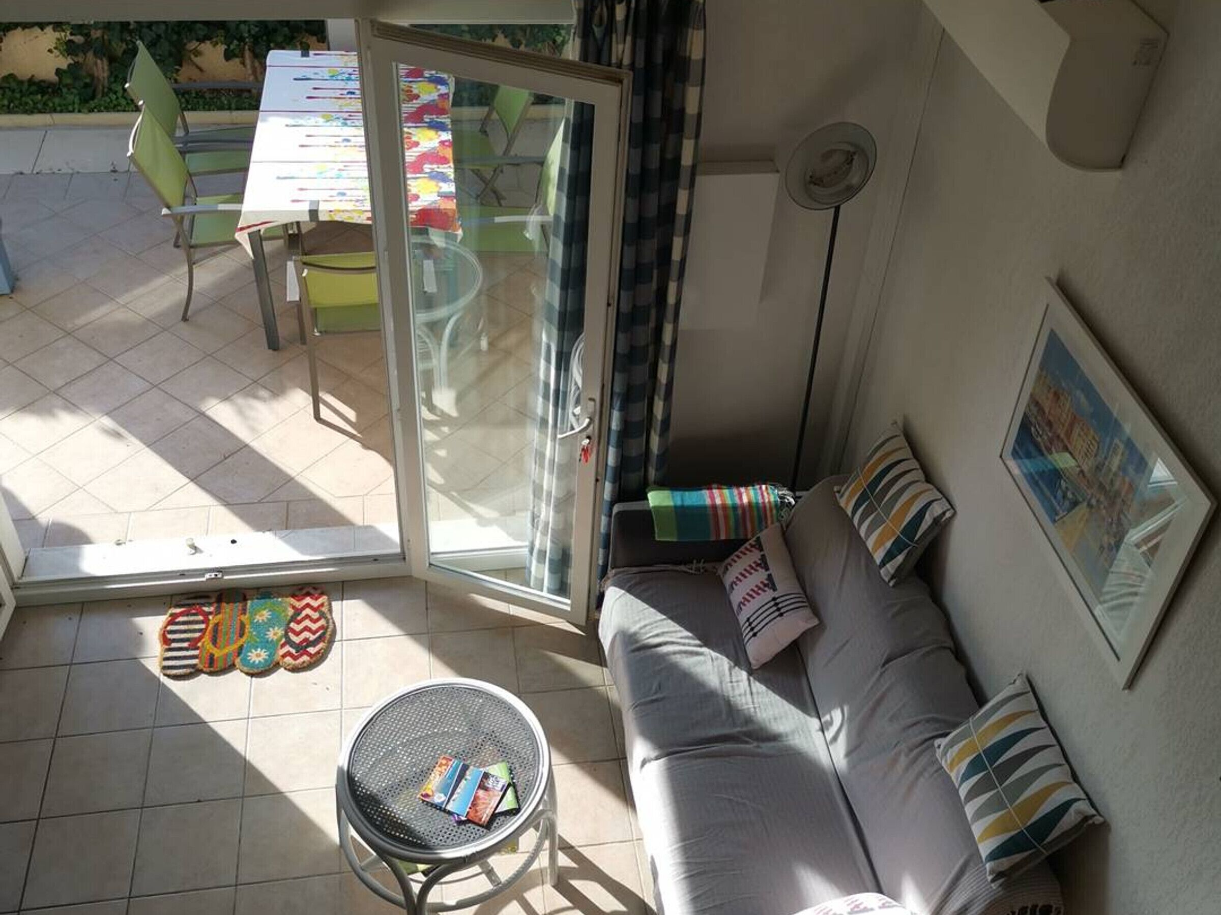 A sunlit room with a sliding glass door, a grey sofa with striped cushions, a round metal table with magazines, and a tiled floor.