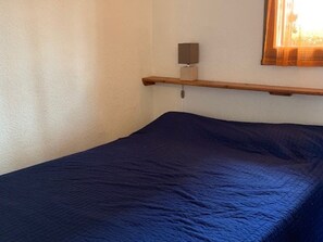 Uma cama de solteiro com colcha azul escuro, mesa de cabeceira com abajur e janela com cortinas.