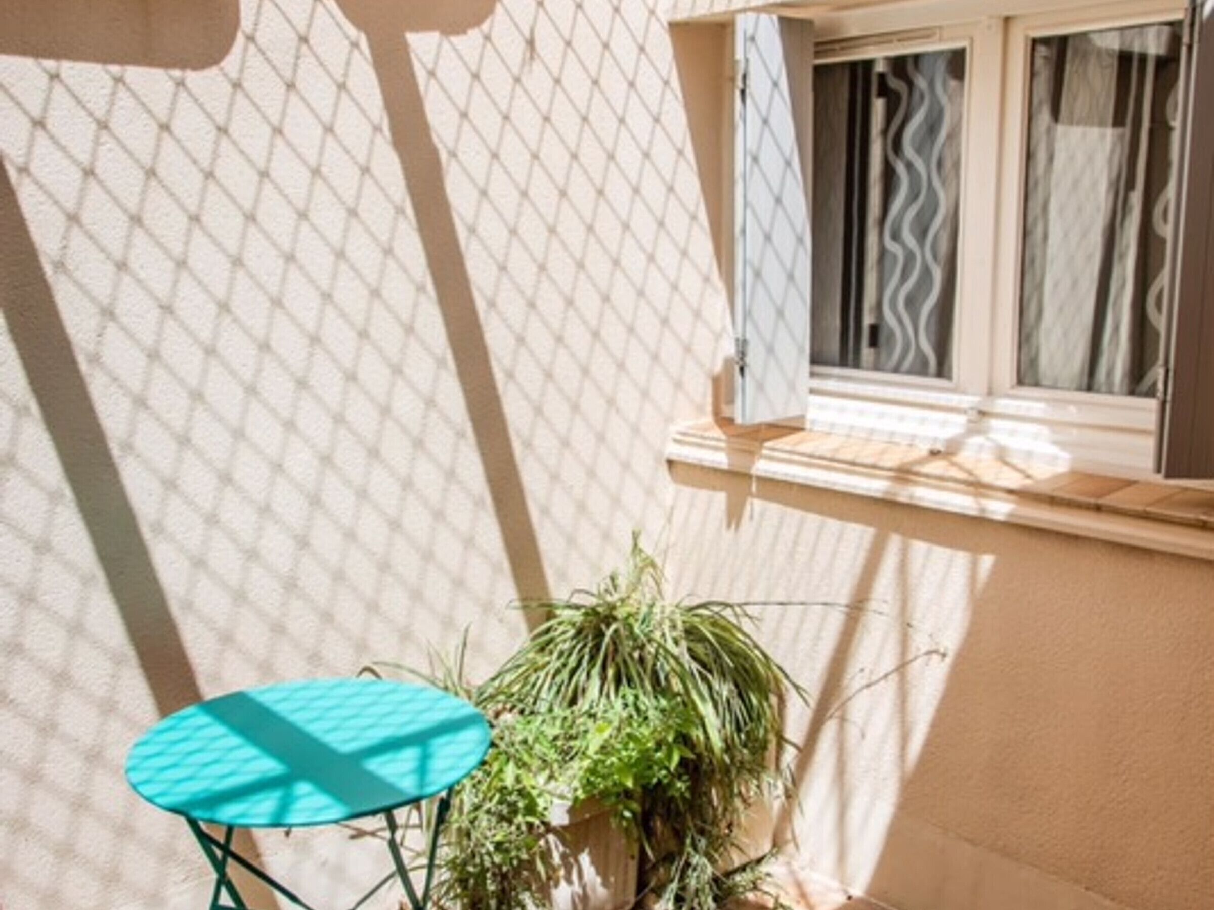 Een klein buitenplaatsje met een turquoise tafel, een plant in pot en een raam met gordijnen.
