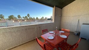 A covered outdoor dining area with a table set for four, chairs, and a view of palm trees and buildings in the background. - Stylish 2BR Apt with Pool, AC & Terrace - Sleeps 6, 600m from Beach (Valras-Plage)