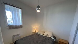 A small, clean bedroom with a single bed, a bedside table with a lamp, a window with curtains, and a radiator. - Stylish 2BR Apt with Pool, AC & Terrace - Sleeps 6, 600m from Beach (Valras-Plage)