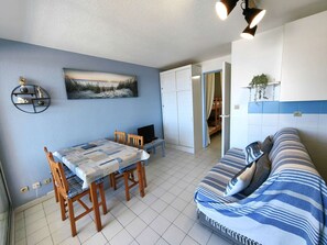 A compact living space with a dining area, a sofa, a TV, and a painting on the wall. - Air-Conditioned T2 Near Beach with Pool and Parking (Cap d'Agde)