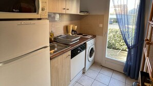 A compact kitchen with a microwave, dishwasher, and washing machine. There is a window with a curtain and a view of greenery outside. - Cozy 3-Room Pavilion with Pool, Near Beach and Shops, Sleeps 6 (Valras-Plage)