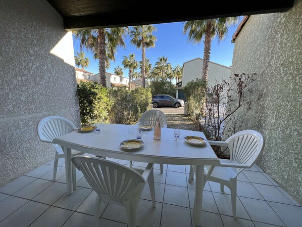 A covered patio with a white table set for two, surrounded by white plastic chairs.