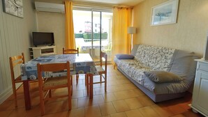 A living room with a sofa, a dining table with chairs, a television, and a balcony with yellow curtains. - Charming A/C T2 with pool and parking, 20m from the sea, sleeps 5 (Cap d'Agde)